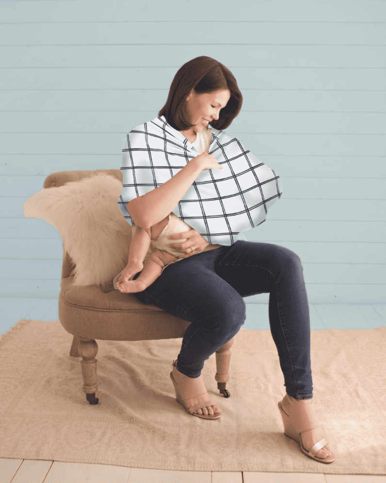 A woman is sitting on a chair holding the Mom Boss™ 4-IN-1 Multi-Use Nursing Cover & Scarf.