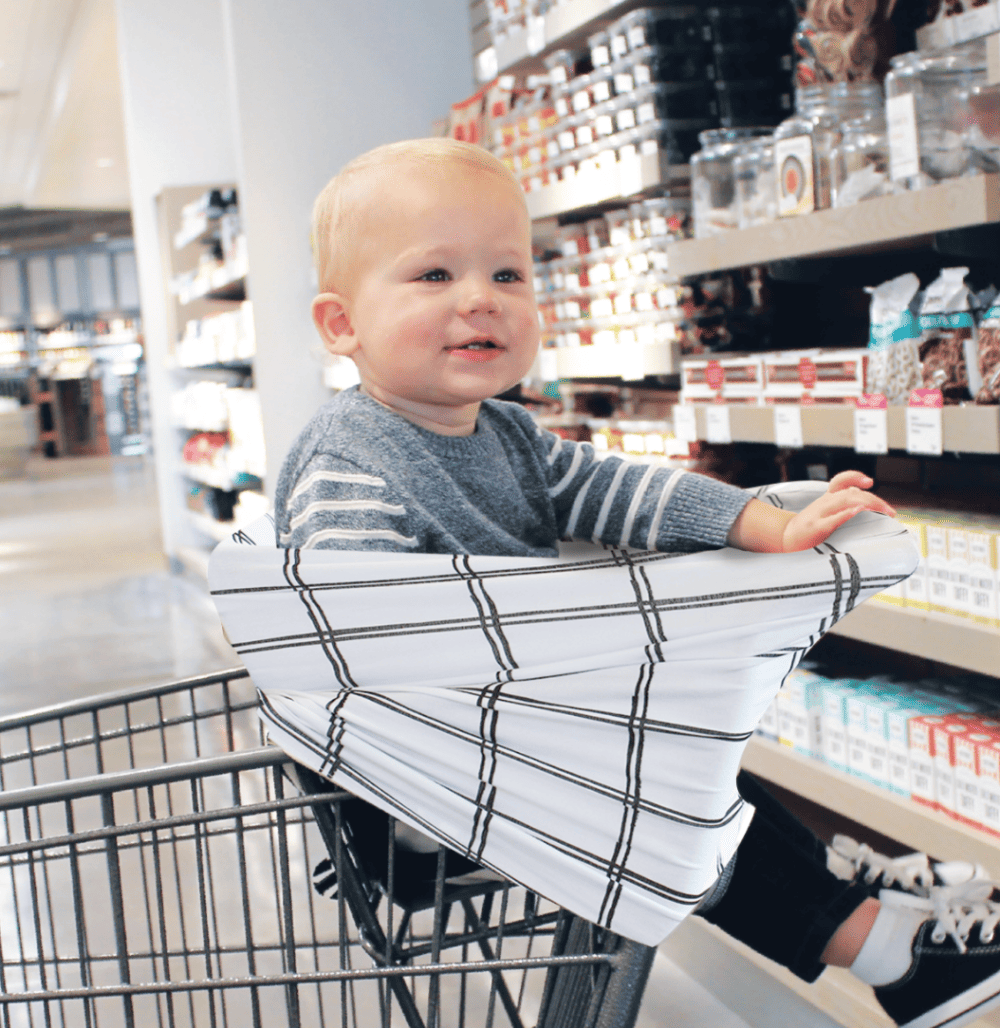 A baby sitting in a Mom Boss™ 4-IN-1 Multi-Use Nursing Cover & Scarf in a grocery store.