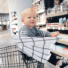 A baby sitting in a Mom Boss™ 4-IN-1 Multi-Use Nursing Cover & Scarf in a grocery store.