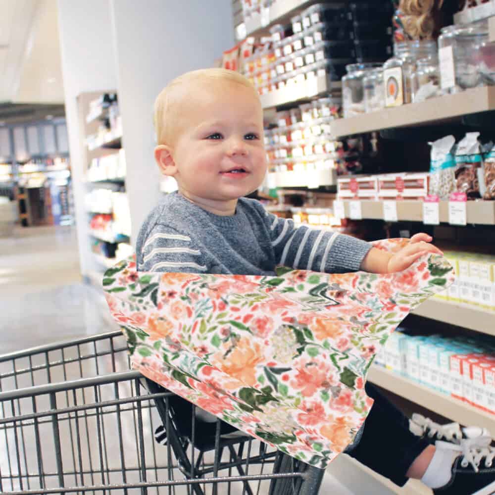 A baby sitting in a Mom Boss™ 4-IN-1 Multi-Use Nursing Cover & Scarf in a grocery store.