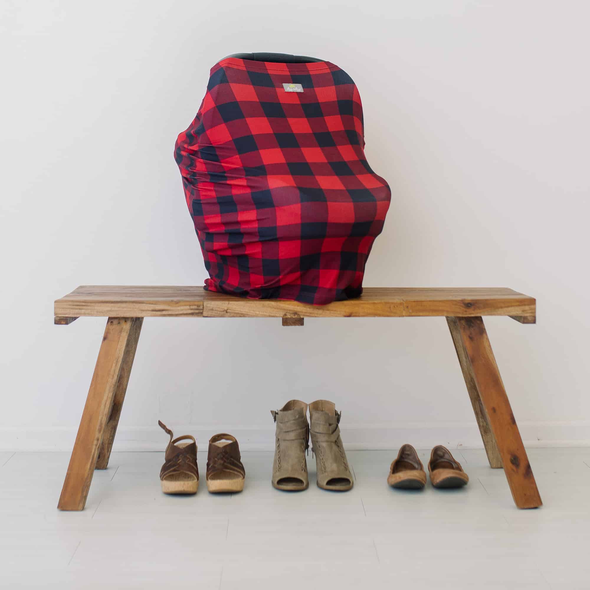 A red and black plaid Mom Boss™ 4-IN-1 Multi-Use Nursing Cover & Scarf on a wooden bench.