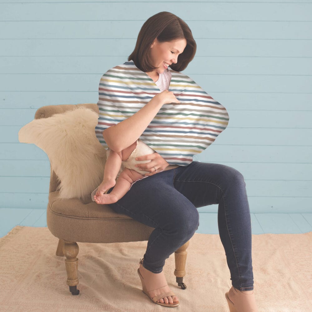 A woman is sitting on a chair with a Mom Boss™ 4-IN-1 Multi-Use Nursing Cover & Scarf in her arms.