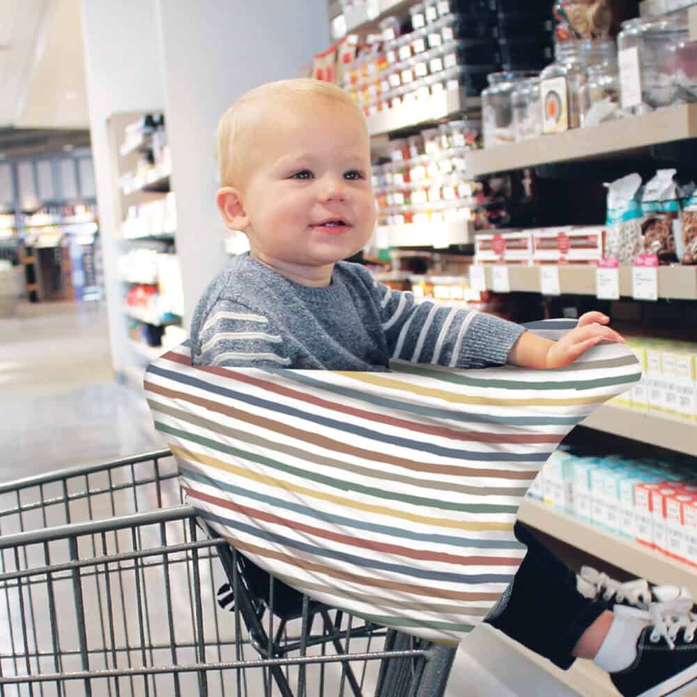 A baby sitting in a Mom Boss™ 4-IN-1 Multi-Use Nursing Cover & Scarf in a grocery store.