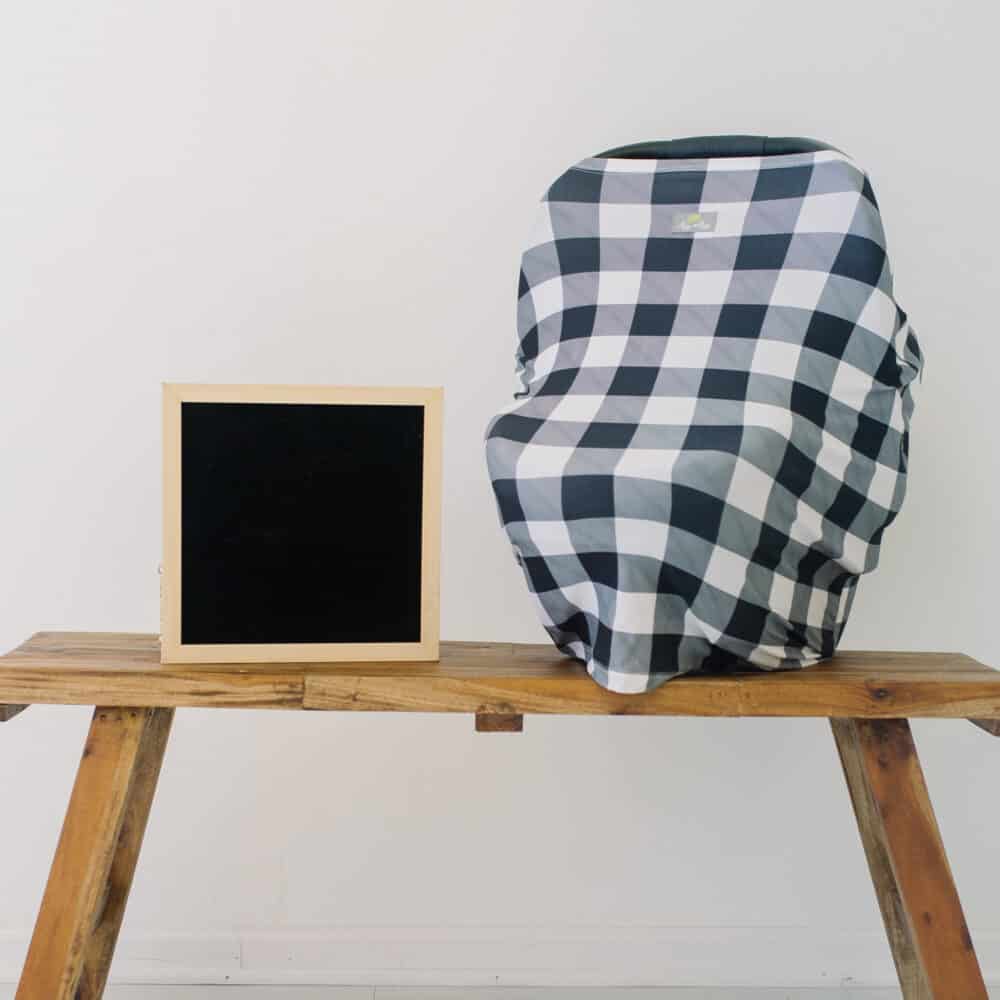 A black and white plaid Mom Boss™ 4-IN-1 Multi-Use Nursing Cover & Scarf on a wooden bench.