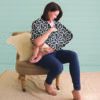 A woman sitting on a chair with a Mom Boss™ 4-IN-1 Multi-Use Nursing Cover & Scarf wrap.