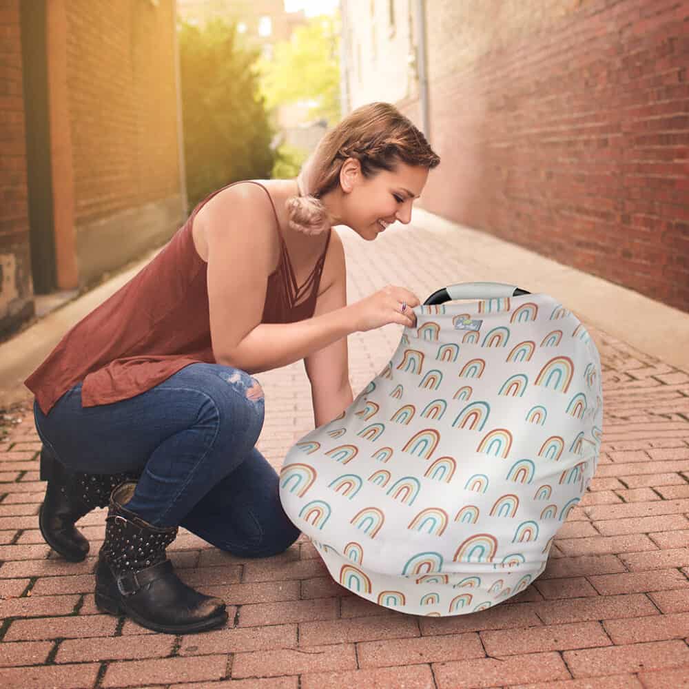 A woman kneeling down on a sidewalk with a Mom Boss™ 4-IN-1 Multi-Use Nursing Cover & Scarf car seat cover.