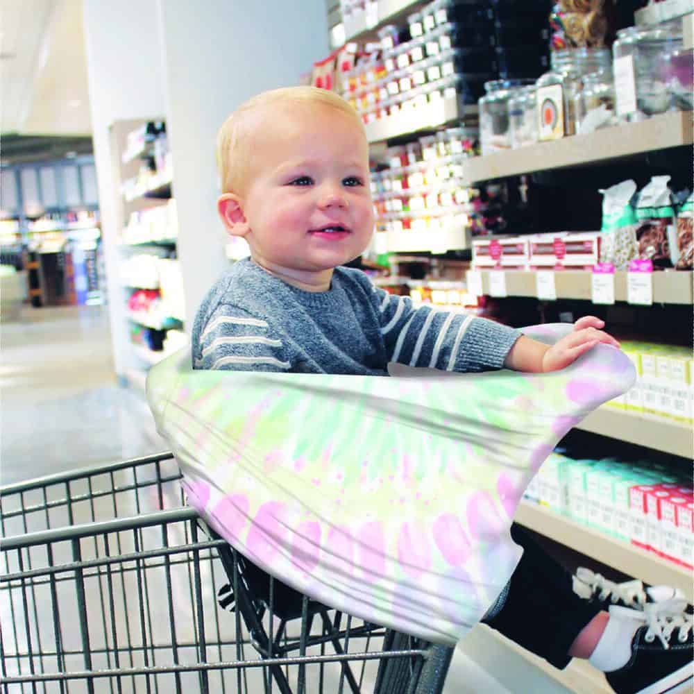 A baby sitting in a Mom Boss™ 4-IN-1 Multi-Use Nursing Cover & Scarf in a grocery store.