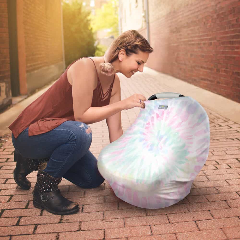 A woman kneeling down next to a Mom Boss™ 4-IN-1 Multi-Use Nursing Cover & Scarf car seat cover.
