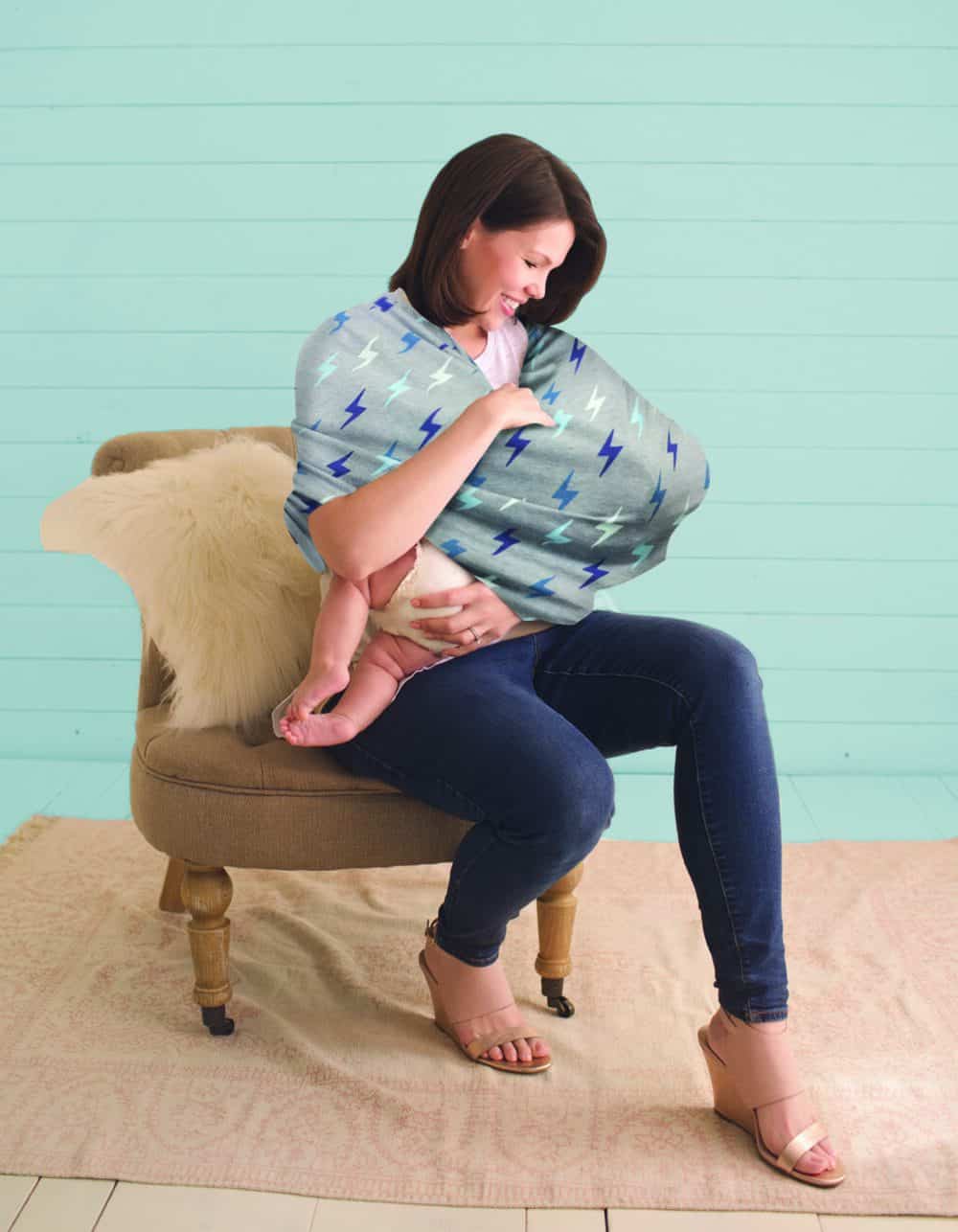 A woman sitting on a chair holding a baby in a Mom Boss™ 4-IN-1 Multi-Use Nursing Cover & Scarf.