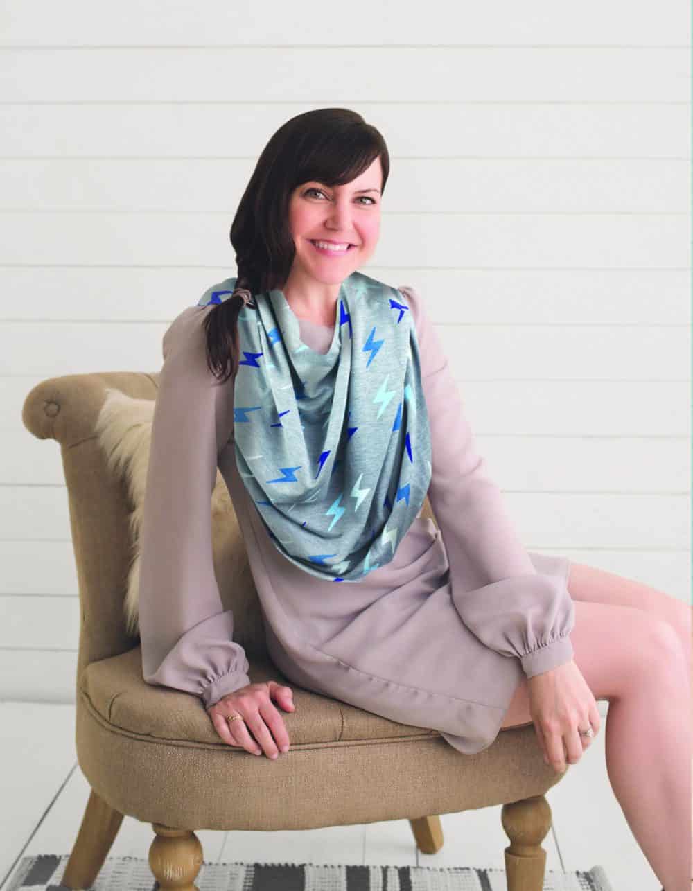 A woman sitting on a chair wearing the Mom Boss™ 4-IN-1 Multi-Use Nursing Cover & Scarf.