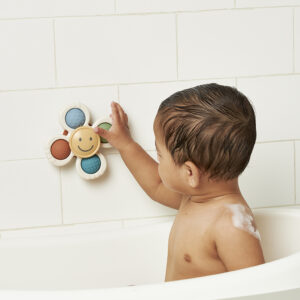 A baby playing with a flower-shaped bath toy attached to a tiled bathroom wall.