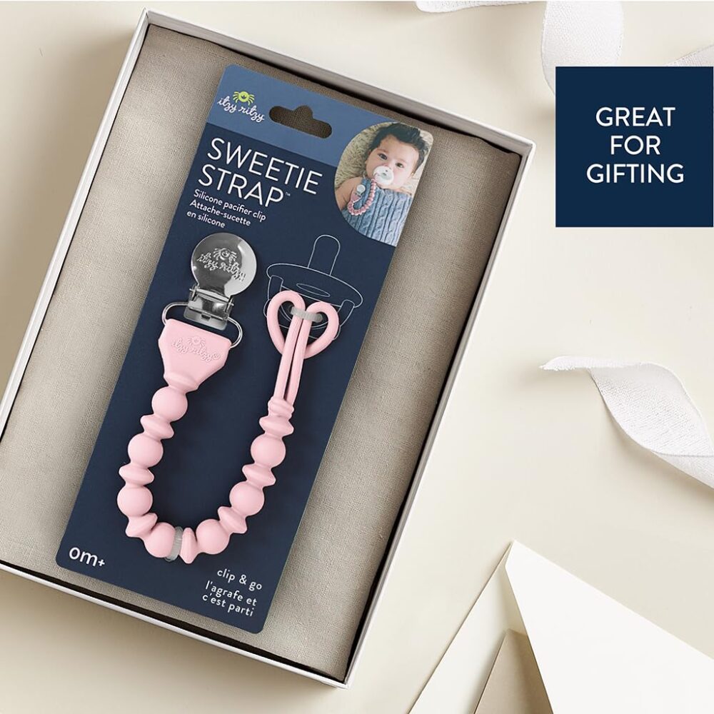 A packaged baby pacifier clip on a beige surface decorated with ribbons. The packaging text reads "Sweetie Strap" and "Great for Gifting" with an image of a baby on it.