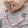 A baby with a pink pacifier clip attached to their gray outfit holds a pacifier in their mouth.