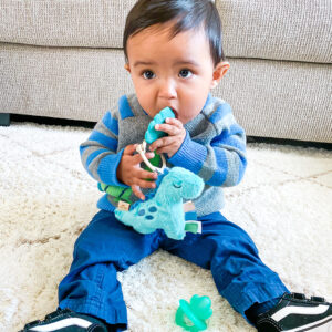 Baby sitting on a white rug, chewing on an Itzy Ritzy - Itzy Pal - Plush Pal Infant Toy with Silicone Teether, wearing a striped sweater and jeans, with a pacifier nearby.
