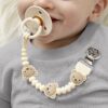 itzy-strap-plus-bear-02 Baby with a pacifier in mouth wears a gray shirt and a pacifier clip featuring beige beads shaped like bear faces.
