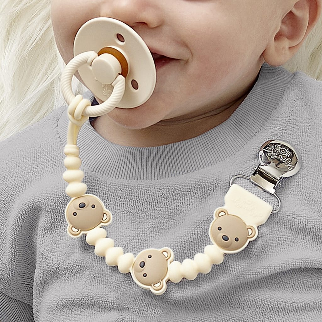 itzy-strap-plus-bear-02 Baby with a pacifier in mouth wears a gray shirt and a pacifier clip featuring beige beads shaped like bear faces.