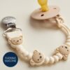 itzy-strap-plus-bear-04 A beige silicone pacifier with a bear-themed beaded clip and a metal clasp lies on a neutral surface. A label reads “FLEXIBLE SILICONE.”.