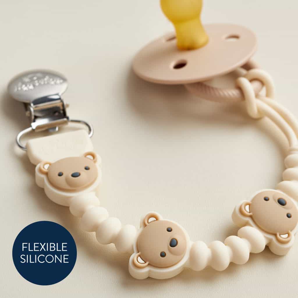 itzy-strap-plus-bear-04 A beige silicone pacifier with a bear-themed beaded clip and a metal clasp lies on a neutral surface. A label reads “FLEXIBLE SILICONE.”.