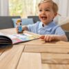 melissadoug-stickerwow-1 A smiling child in a blue shirt sits at a wooden table, holding the Melissa & Doug Sticker WOW Activity Pad and excitedly pointing at its colorful, open pages.