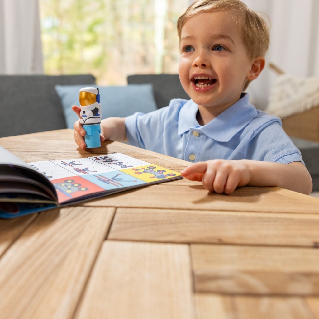 melissadoug-stickerwow-1 A smiling child in a blue shirt sits at a wooden table, holding the Melissa & Doug Sticker WOW Activity Pad and excitedly pointing at its colorful, open pages.