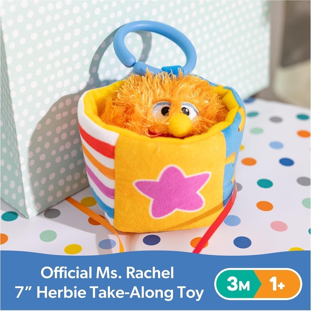 A plush orange toy with a blue handle sits in a yellow, star-decorated box on a polka dot surface. Text reads: “Ms Rachel Official Sensory Take-Along Toy Herbie Doll – Baby Teether, Rattle, Stroller Clip, 3 Months+.”.