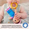 A baby chews on the yellow teether attached to the Ms Rachel Official Sensory Take-Along Toy Herbie Doll while holding it with both hands. Text at the bottom highlights the toy’s sensory features, including its rattle and convenient stroller clip for babies 3 months and up.