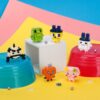 nanoblock-tamagotchi-135 Six vibrant, pixelated Tamagotchi figures from the Kawada nanoblock mininano Blind Box First Generation set are displayed on plastic bowls and a cube, with a bright yellow and blue backdrop and scattered small beads.