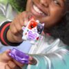 nanomals-119 A child holds up the Hasbro Nano Mals Purrnelope Calico Kitty Electronic Pet Interactive Fidget Toy Nanomals figure, featuring a purple heart on its chest and a matching purple accessory, smiling in the background.