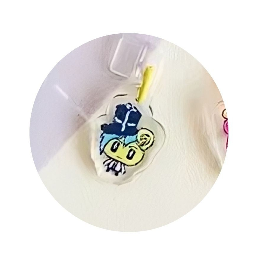 tamagotchi-uni-dustplug-3 A Tamagotchi Uni keychain dust plug featuring a cartoon animal face with a blue hat and bowtie, attached to a clear acrylic charm for mobile phones with Type C ports.